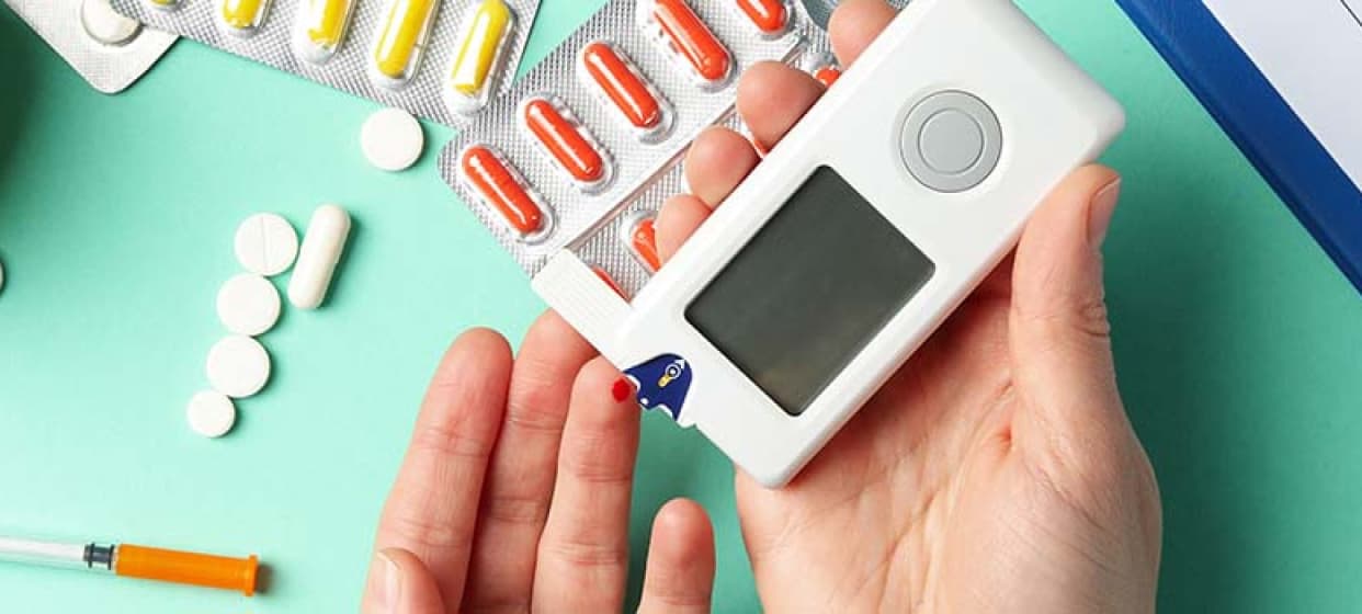 An image showing hands with a diabetes monitoring device and some pills placed on a table.