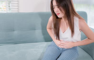 An image of a woman sitting on her couch, holding her stomach with a pained expression.